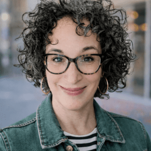 Person with short, curly dark hair wearing large hoop earrings, a green denim jacket, and a black-and-white striped shirt, standing outdoors with a blurred background.