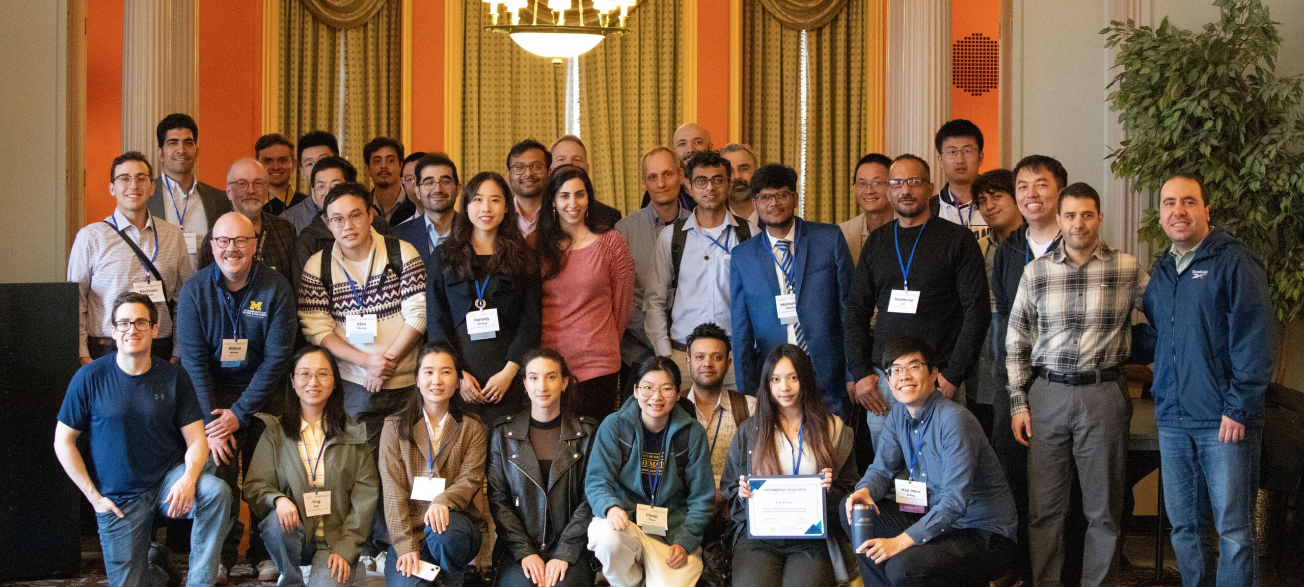 A large group poses in a university room after the conference, wearing name tags and one is holding a certificate.