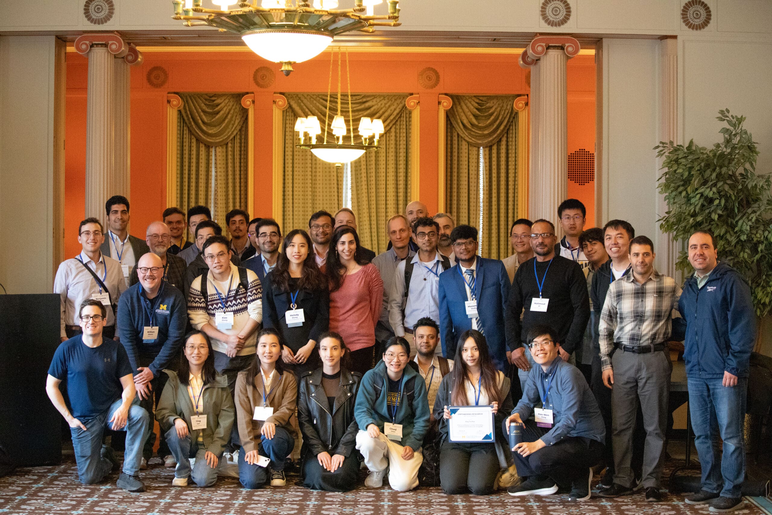 A large group poses in a university room after the conference, wearing name tags and one is holding a certificate.