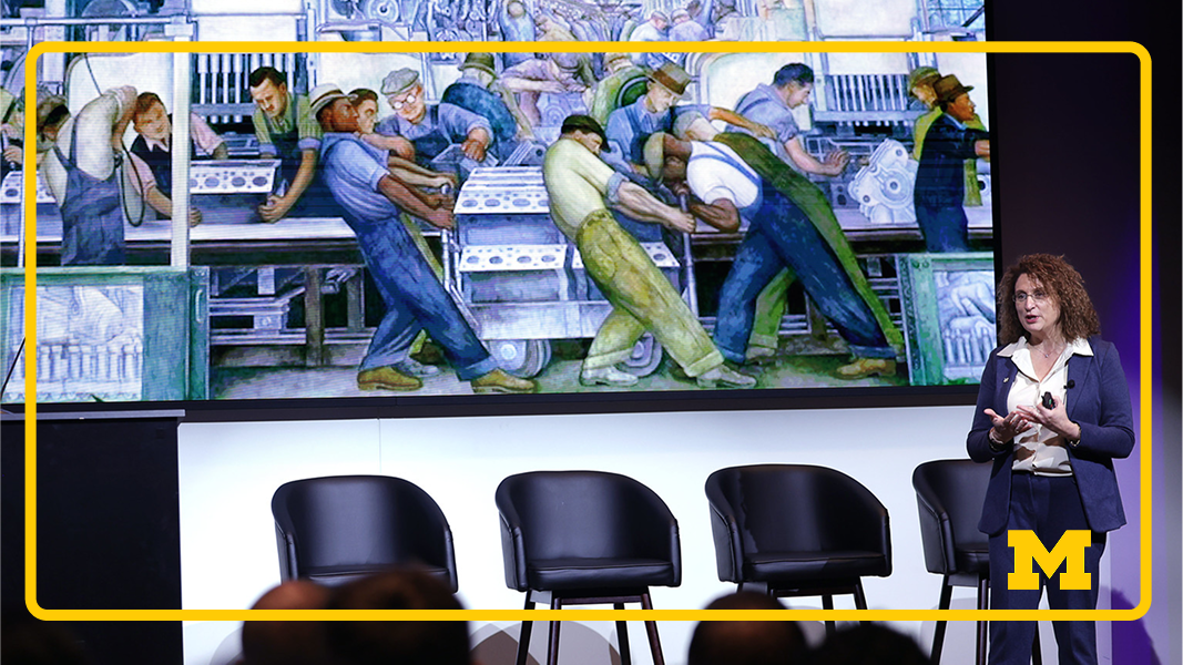A woman speaks before a crowd with a drawing behind her of workers.